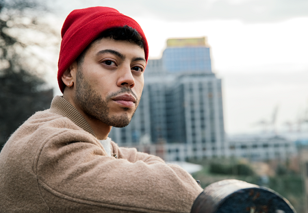 A man wearing a red beanie stares into the camera with a high rise building in the background