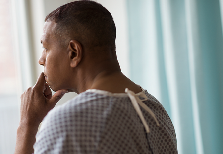 A patient wearing a hospital gown stoically looks out the window, lost in thought
