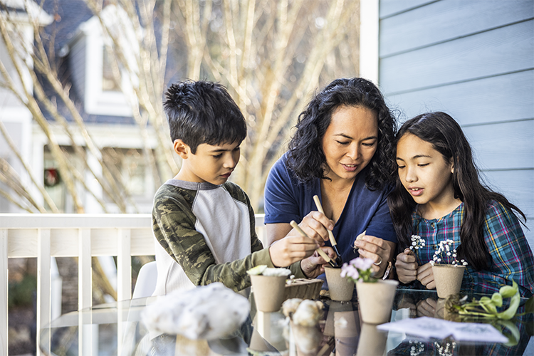 A Native American mother and her 2 school age children planting flowers at a table on a front porch