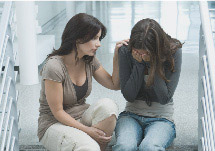 Two young women. One crying with her hands over her face, the other with her hand on the shoulder of the crying woman.