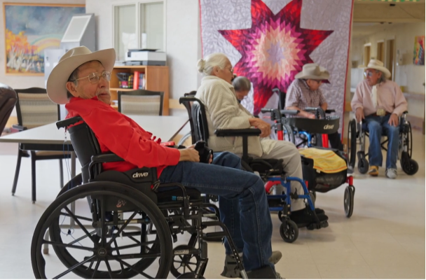 Five elders in wheelchairs congregate near a wall decorated with a quilt.