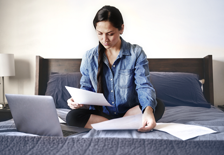 A woman sitting on her bad with an open laptop in front of her as she looks at the papers in her hands