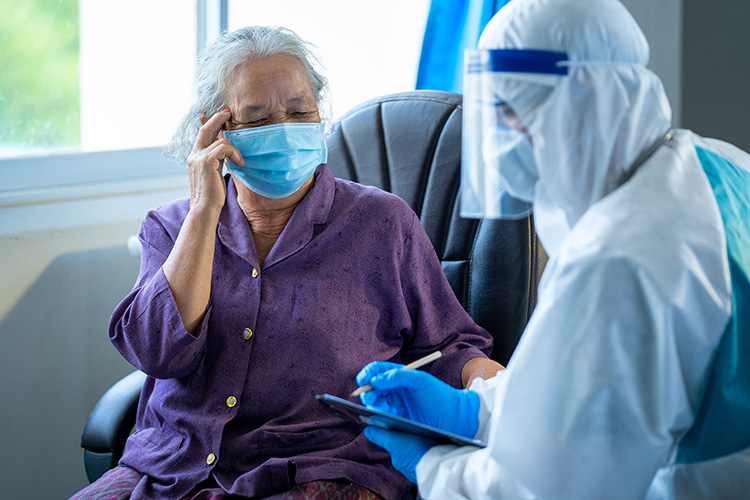 A medical person covered in white from head to toe, wearing a mask, face shield, and gloves taking notes while talking to an elderly woman wearing a mask.