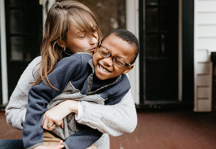 A young white woman smiling and hugging a young, smiling, black boy with glasses