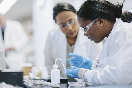 Two female scientists wearing white lab coats and goggles working at the bench