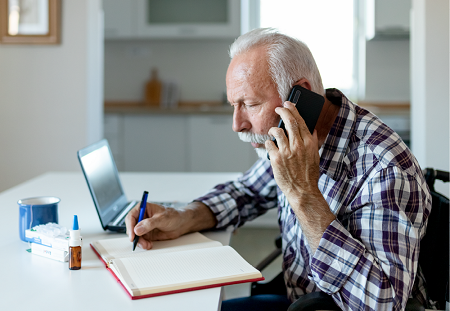 A seated elderly man with a laptop and medicine in front of takes notes as he talks on the phone