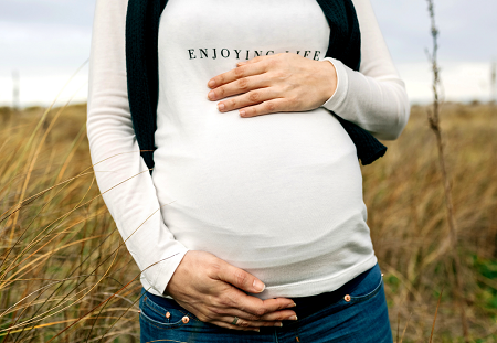 Close up of the belly of a pregnant woman standing in a field, her shirt reads "enjoying life"