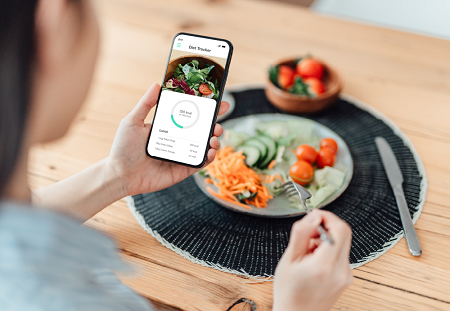 Vantage point over the shoulder of a woman as she looks at her diet tracker app on her phone with a plate of vegetables in front of her