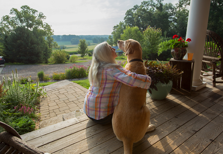 Seated woman on the edge of her porch has her arm around her dog with a countryside landscape in the background